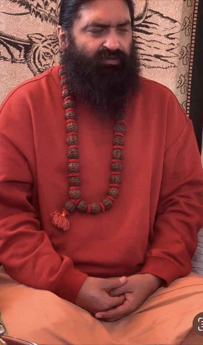 Swami Aditya seated in quiet meditation, eyes lowered and hands folded in his lap, wearing a rudraksha mala