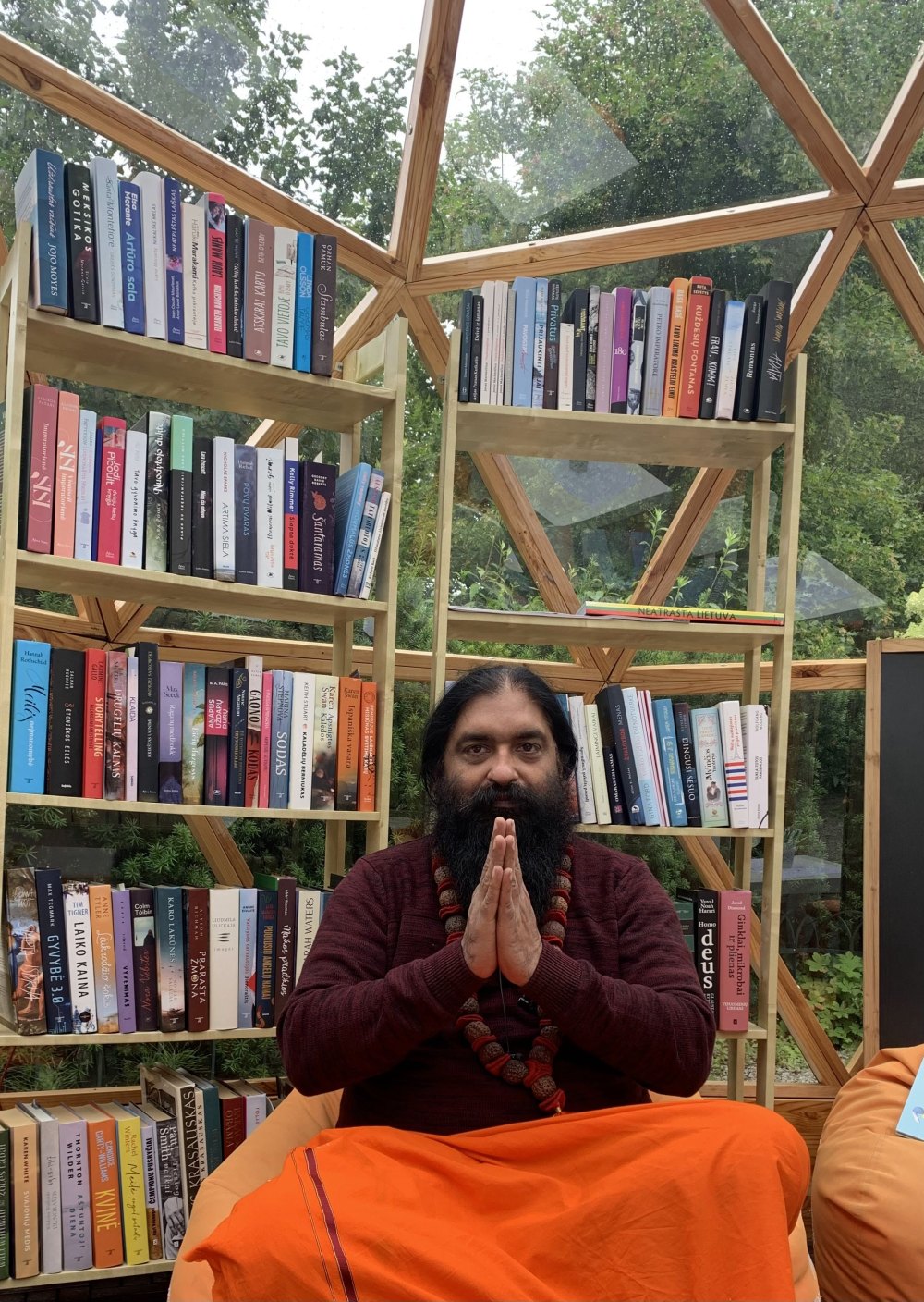 Swami Aditya seated in a glass garden dome lined with books, hands joined in namaste