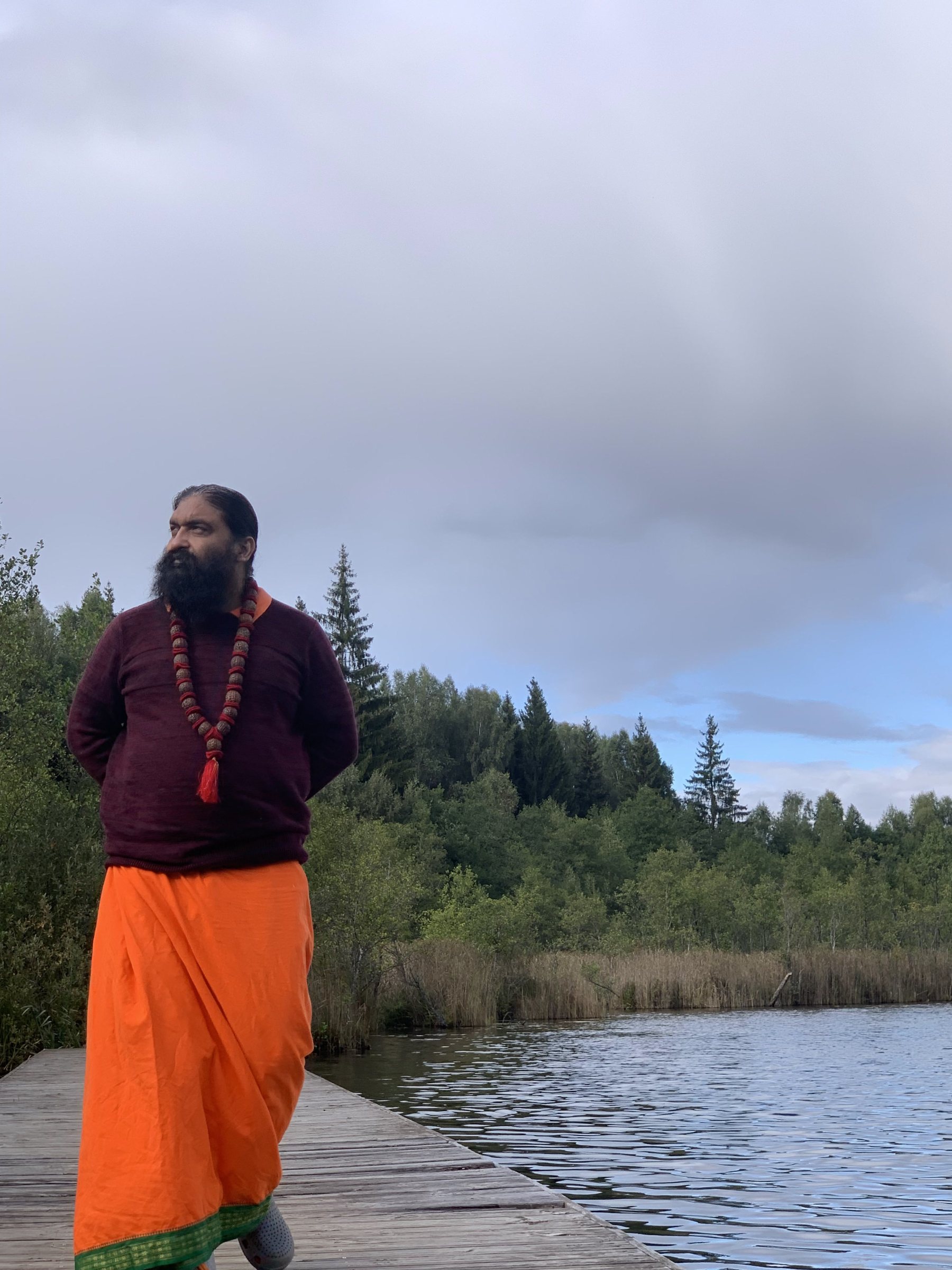 Swami Aditya standing at the edge of a still northern lake under an open sky