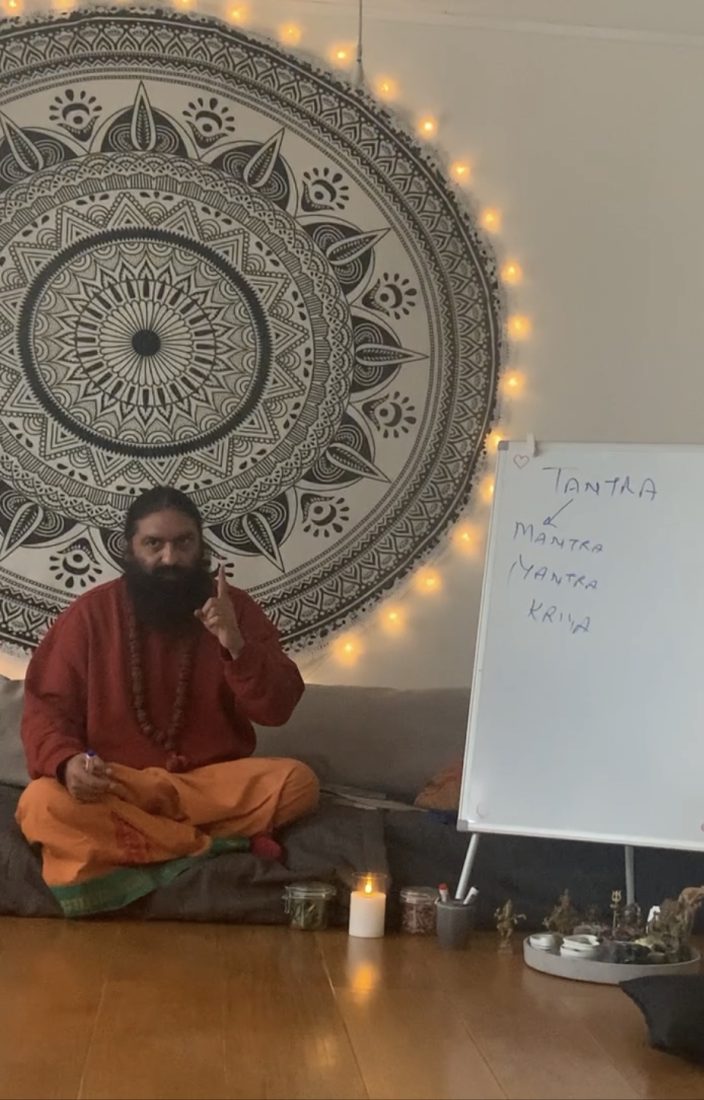 Swami Aditya seated cross-legged before a mandala tapestry, finger raised in teaching beside a whiteboard reading tantra, mantra, yantra, kriya